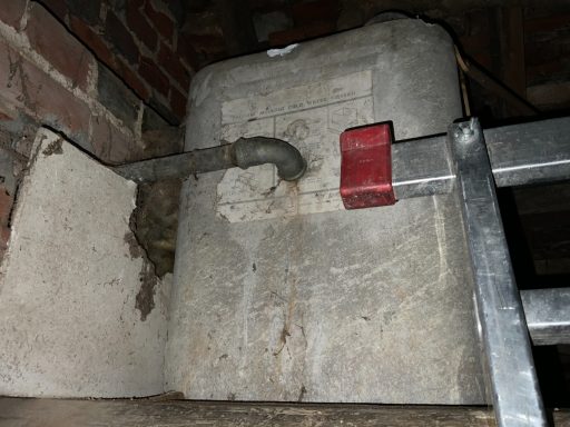 Old grey water tank with a pipe attached, positioned against a brick wall.